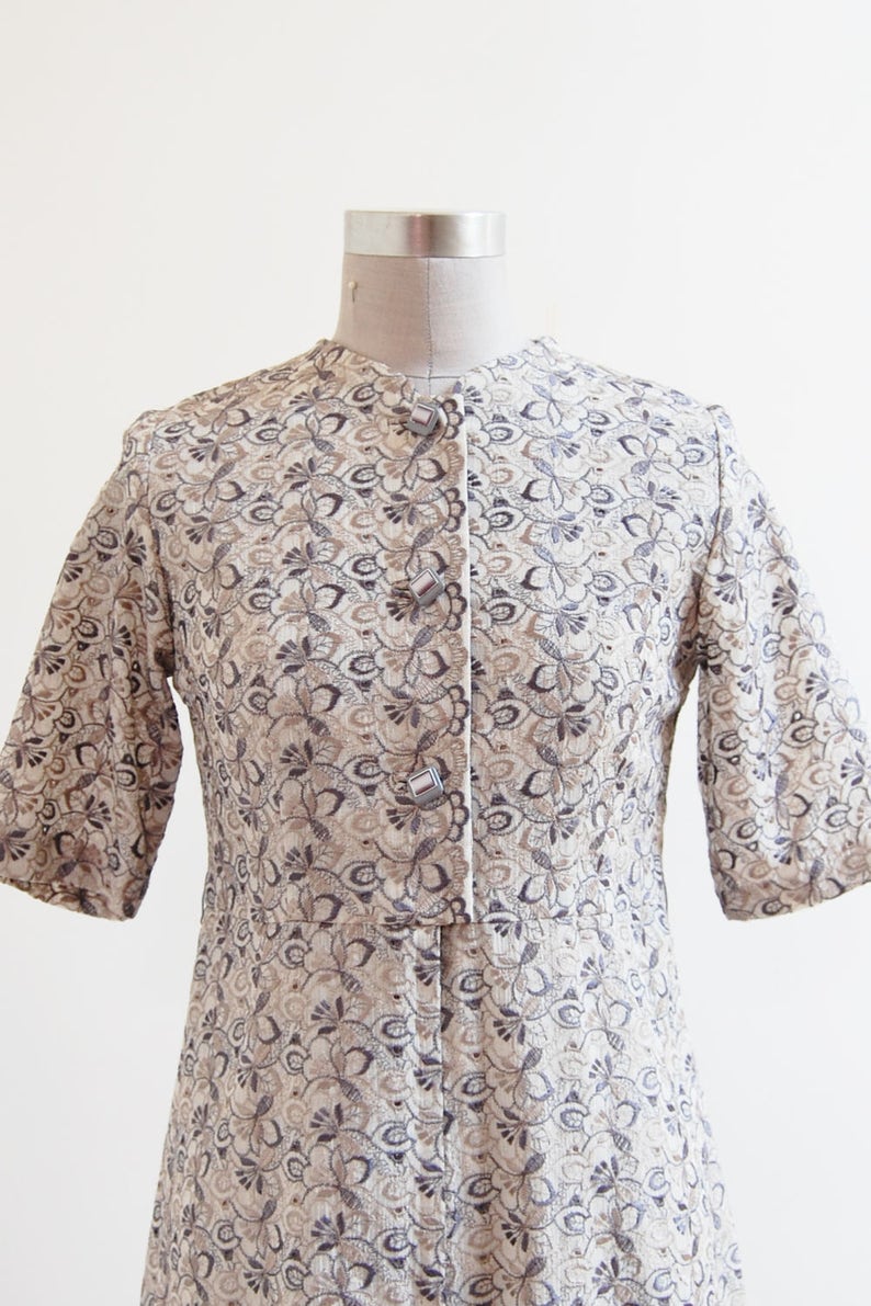 May include: A beige dress with a floral pattern and silver buttons. The dress has a short sleeve and a fitted bodice.