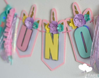 May include: A pink, yellow, and blue unicorn-themed banner with the word "UNO" spelled out. The banner is decorated with flowers, glitter, and ribbons.