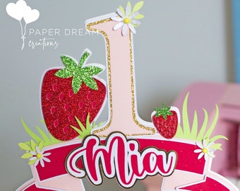 May include: A pink and red cake topper with a large number one, a strawberry, and the name "Mia" written in a pink banner. The topper is decorated with glitter and small white flowers.