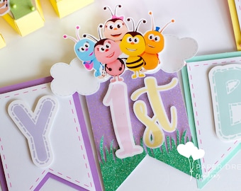 May include: A purple, green, and yellow banner with the word "1st" in the center. The banner is decorated with a cloud and five cartoon bees. The banner is made of paper and has a glittery finish.