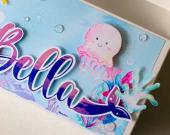 May include: A light blue and white gift box with the words "PAPER DREAM creations" on the front. The box is decorated with a pink and purple seashell, a pink jellyfish with a smiling face, and the name "Bella" in a colorful, stylized font.