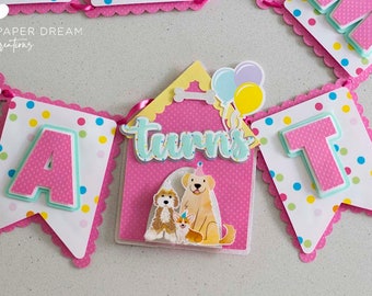 May include: A pink and white banner with the words "A turns T" and a dog house with three dogs inside. The banner has colorful polka dots and a scalloped edge.