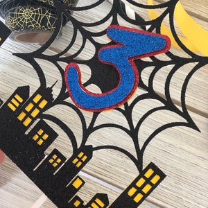 Puede incluir: Topper de pastel de telara&ntilde;a negra y roja con un n&uacute;mero 3 de purpurina azul en el centro. La telara&ntilde;a est&aacute; sobre un paisaje urbano negro con ventanas amarillas.