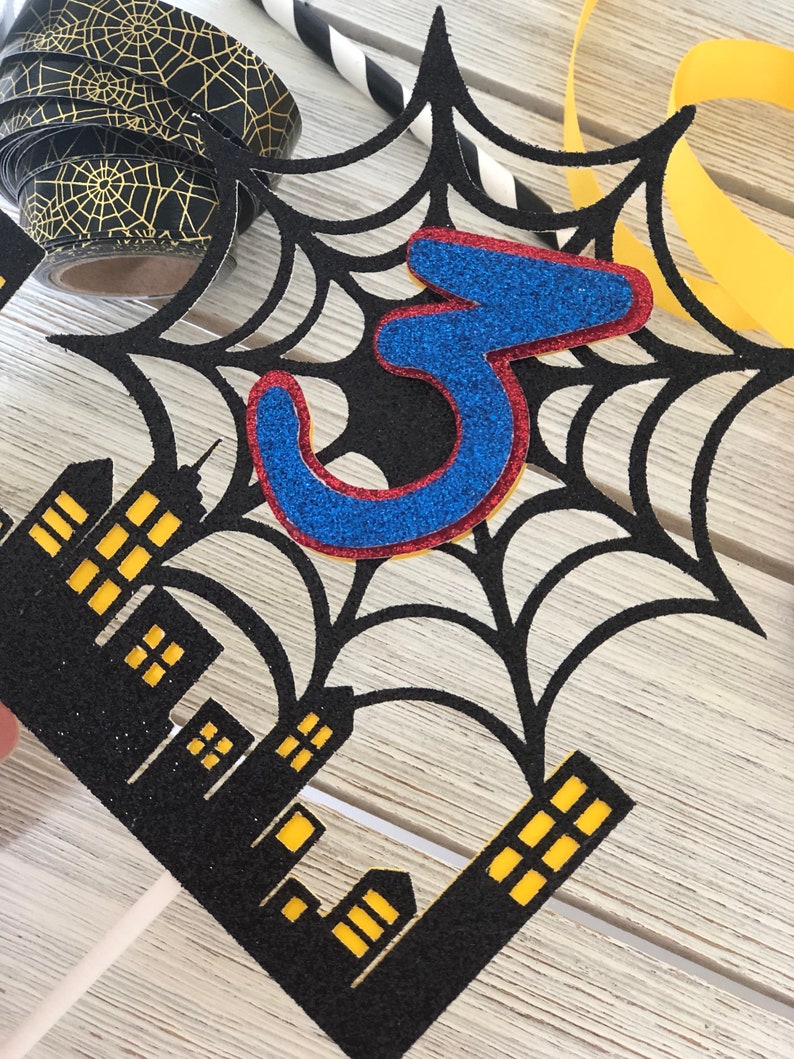 Puede incluir: Topper de pastel de telara&ntilde;a negra y roja con un n&uacute;mero 3 de purpurina azul en el centro. La telara&ntilde;a est&aacute; sobre un paisaje urbano negro con ventanas amarillas.