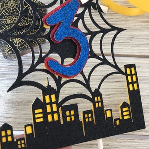 Puede incluir: Topper de pastel de telara&ntilde;a negra y amarilla con un n&uacute;mero 3 de purpurina azul y roja y una ciudad negra con ventanas amarillas.