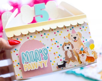 May include: A white and yellow party favor box with a confetti pattern. The box has a scalloped edge and a pink arch with the text "MIA'S PAW-TY". There are four cartoon dogs wearing party hats and a cluster of balloons.