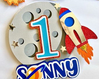 May include: A cake topper featuring a gray moon with a blue number one, a red and gold rocket, and a blue name "SONNY" with a small orange planet. The topper is perfect for a first birthday party with an outer space theme.
