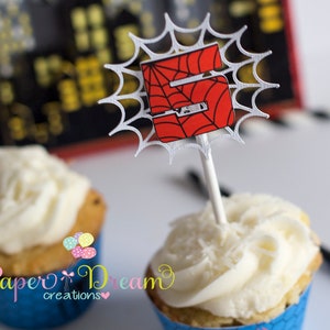 Puede incluir: Un cupcake blanco con glaseado blanco cubierto con un adorno de cupcake de telara&ntilde;a roja y negra de Spiderman. El adorno presenta el n&uacute;mero 5 en el centro de la telara&ntilde;a.