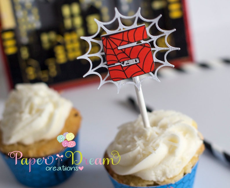 Puede incluir: Un cupcake blanco con glaseado blanco cubierto con un adorno de cupcake de telara&ntilde;a roja y negra de Spiderman. El adorno presenta el n&uacute;mero 5 en el centro de la telara&ntilde;a.