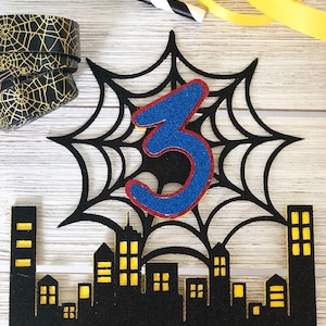 Puede incluir: Topper de pastel negro y amarillo con una telara&ntilde;a negra y un n&uacute;mero 3 azul en el centro. La telara&ntilde;a est&aacute; sobre un paisaje urbano negro con ventanas amarillas.