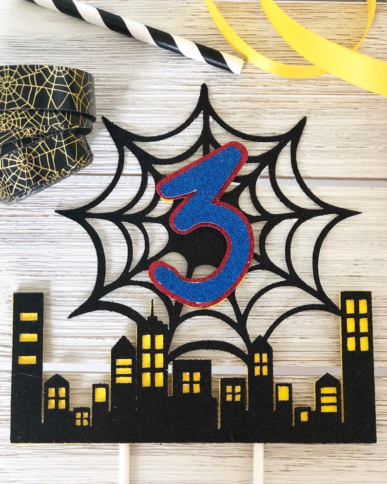 Puede incluir: Topper de pastel negro y amarillo con una telara&ntilde;a negra y un n&uacute;mero 3 azul en el centro. La telara&ntilde;a est&aacute; sobre un paisaje urbano negro con ventanas amarillas.