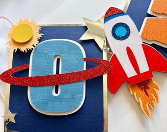 May include: A blue and red rocket ship with a white body and orange flames. The rocket is attached to a blue banner with a white number "0" and a red ring around it. The banner has a silver border and a white star in the corner.
