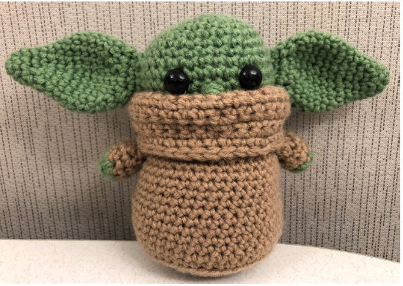 以下が含まれることがあります： スターウォーズのフランチャイズのキャラクターに似ている、かぎ針編みで作ったぬいぐるみ。おもちゃは緑と茶色で、大きな黒い目と茶色の編み物のマフラーをしています。