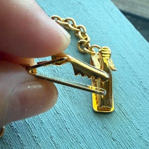 May include: A gold-colored tie clip with a bottle-shaped design. The clip features a chain and a clasp to attach to a tie. The clip is held by a person's fingers against a blue background.