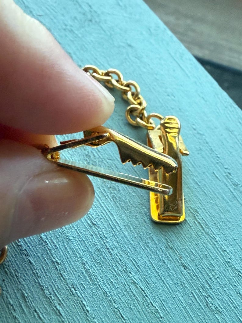 May include: A gold-colored tie clip with a bottle-shaped design. The clip features a chain and a clasp to attach to a tie. The clip is held by a person's fingers against a blue background.