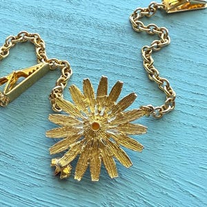 May include: A gold-toned tie clip with a daisy-shaped pendant. The pendant has detailed petals and a small star accent. The clip features a chain and a clasp for securing to a tie. The background is a light blue surface.