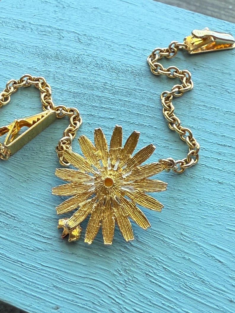 May include: A gold-toned tie clip with a daisy-shaped pendant. The pendant has detailed petals and a small star accent. The clip features a chain and a clasp for securing to a tie. The background is a light blue surface.