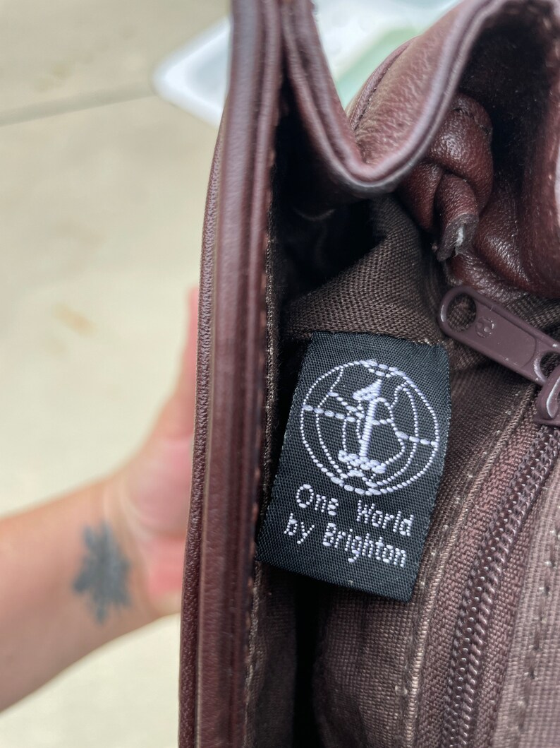 May include: A brown leather purse with a black tag that reads "One World by Brighton".