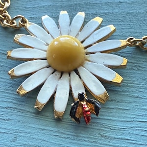 May include: A gold-tone necklace featuring a large daisy pendant with white petals, a yellow center, and gold-colored accents. A small, detailed bee charm with black and red wings is attached to the daisy. The chain is also gold-colored.