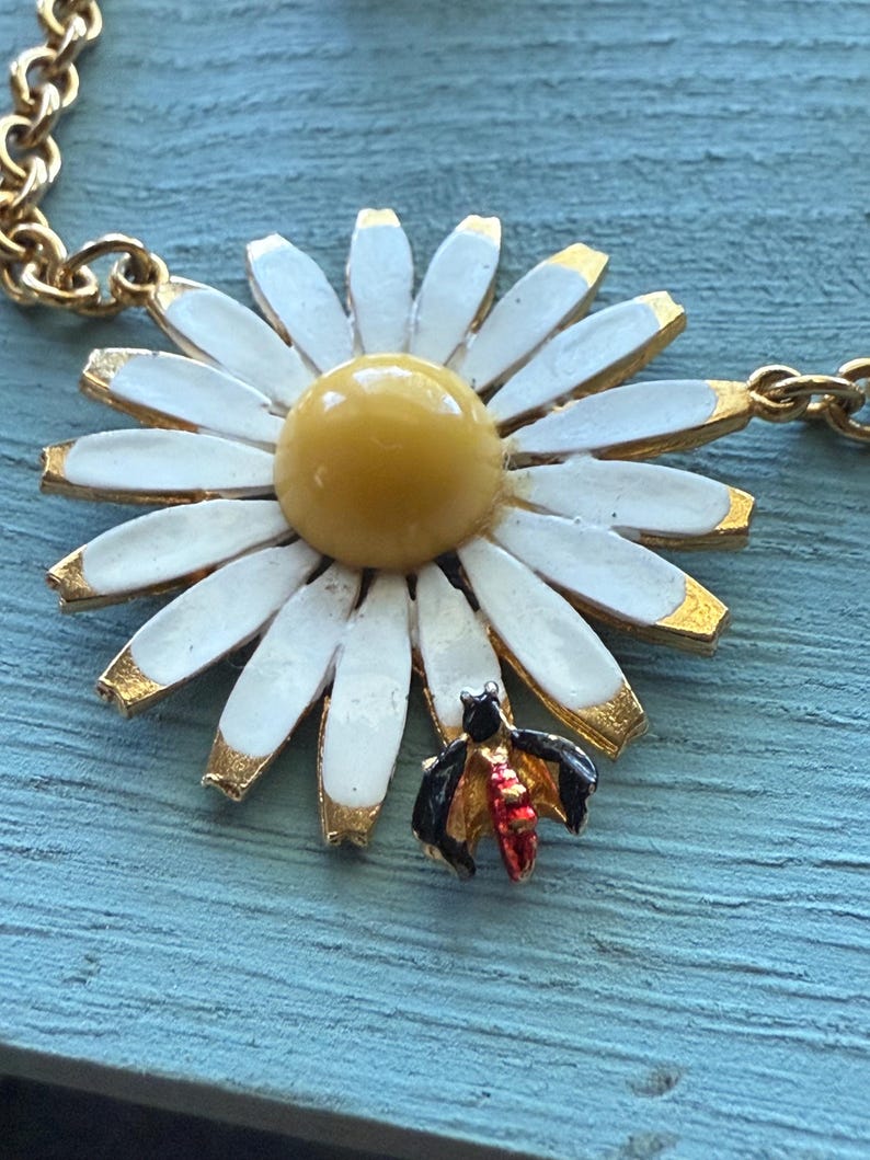 May include: A gold-tone necklace featuring a large daisy pendant with white petals, a yellow center, and gold-colored accents. A small, detailed bee charm with black and red wings is attached to the daisy. The chain is also gold-colored.