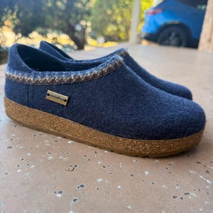 May include: A pair of navy blue felt slippers with a cork sole. The slippers have a decorative stitched trim around the opening and a small gold-colored tag that reads "HAFLINGER". These cozy slip-ons are ideal for indoor wear.