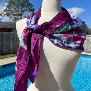 May include: A purple and white floral patterned silk scarf with a green and blue design. The scarf is tied around a white mannequin's neck.