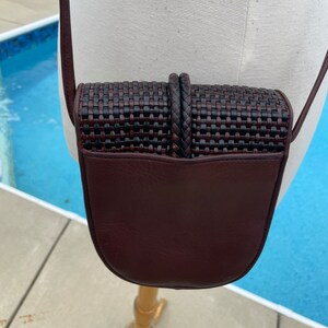 May include: A small brown leather crossbody bag with a woven flap. The bag has a dark brown leather strap and a rounded bottom.