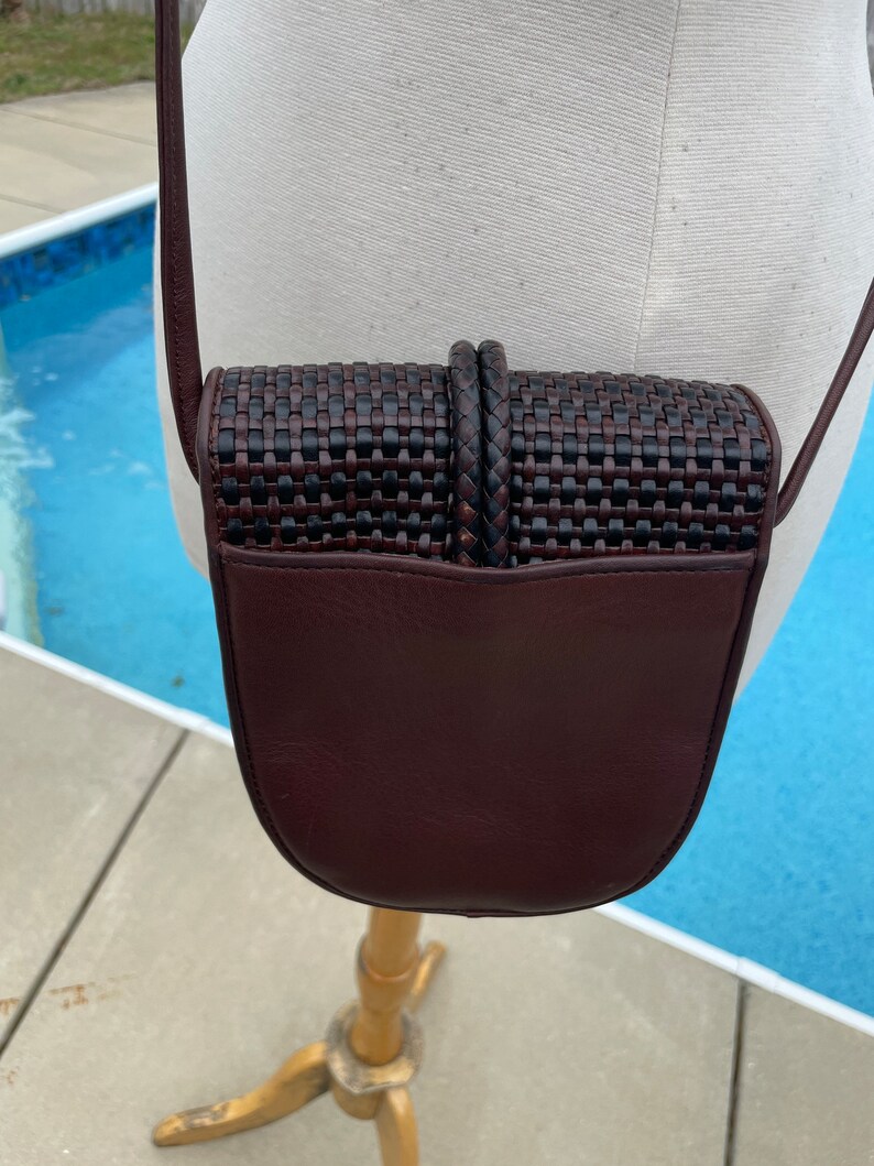 May include: A small brown leather crossbody bag with a woven flap. The bag has a dark brown leather strap and a rounded bottom.