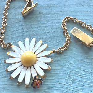 May include: A vintage daisy brooch with a gold chain and clips. The white enamel petals have gold edges, surrounding a yellow center. A small bee charm with red and black details is attached. The chain and clips are gold-toned.