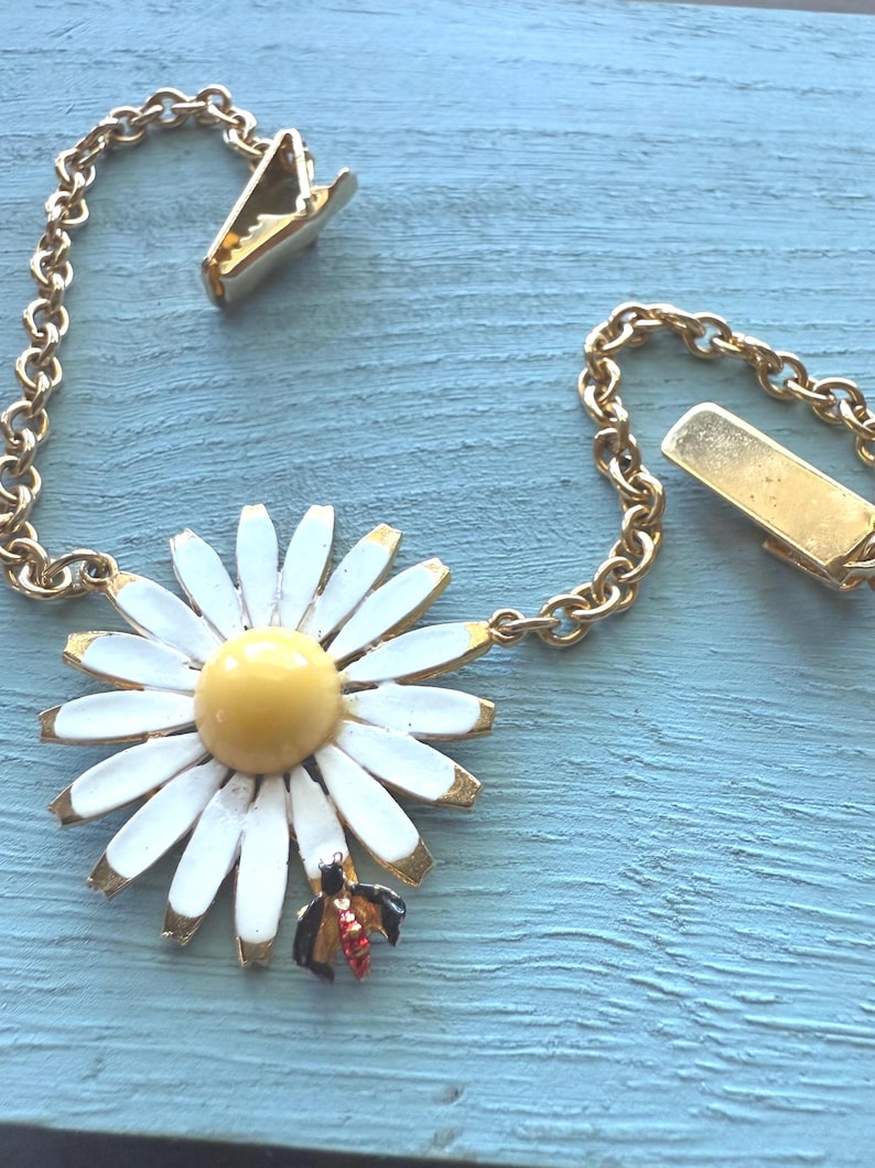 May include: A vintage daisy brooch with a gold chain and clips. The white enamel petals have gold edges, surrounding a yellow center. A small bee charm with red and black details is attached. The chain and clips are gold-toned.