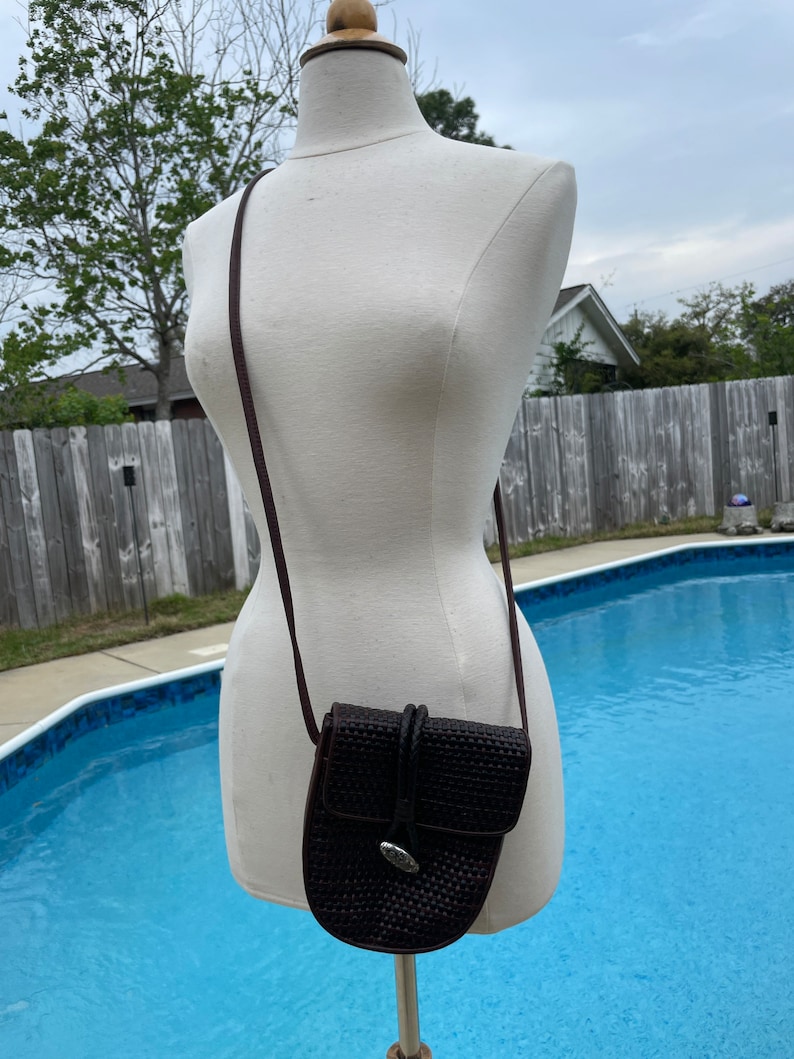 May include: A small brown woven leather crossbody bag with a silver buckle. The bag is being modeled on a white mannequin against a backdrop of a swimming pool and a wooden fence.
