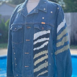 May include: A blue denim jacket with silver stars and a white and black striped design on the front and a green and white striped design on the right sleeve. The jacket has a brown leather label that says "RUSTLER".