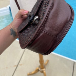 May include: A brown leather and woven crossbody bag with a silver clasp. The bag has a flap closure and a woven strap.