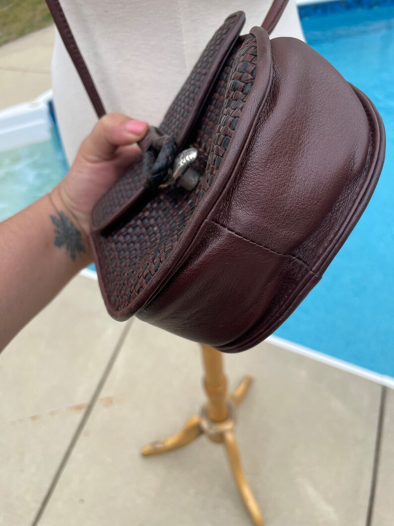 May include: A brown leather and woven crossbody bag with a silver clasp. The bag has a flap closure and a woven strap.