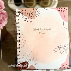 May include: A spiral-bound planner with a soft pink and white watercolor design. The cover features floral and abstract shapes in shades of brown and pink, with the text "School Psychologist Planner". The planner is displayed on a clear stand.
