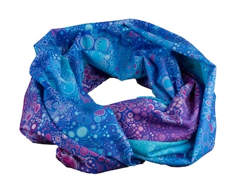 May include: A blue infinity scarf with a repeating pattern of circles in shades of blue, teal, and purple. The fabric is soft and appears to be lightweight, suitable for various weather conditions. The scarf is a tube shape.