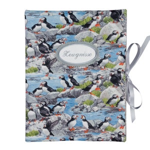 Könnte beinhalten: Ein graues, stoffbezogenes Tagebuch mit einem blauen und weißen Muster aus Papageientauchern auf Felsen. Das Tagebuch hat ein graues Band, das um es gebunden ist, und ein graues ovales Etikett mit der Aufschrift "Zeugnisse".