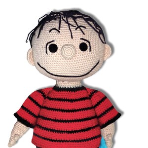 Puede incluir: Un juguete de peluche tejido a crochet de un niño con cabello negro, que lleva una camisa a rayas rojas y negras, pantalones cortos negros y zapatos marrones. El juguete sostiene un paño azul claro.