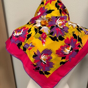 May include: A square scarf with a vibrant floral pattern. The scarf features large flowers in shades of purple, pink, and white against a bright yellow background. A hot pink border frames the design. The scarf is displayed on a mannequin.