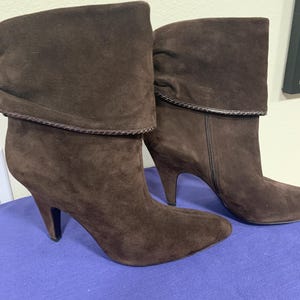 May include: A pair of brown suede ankle boots with a folded-over cuff design. The boots have pointed toes, stiletto heels, and a side zipper. The heels are approximately 3 inches high.