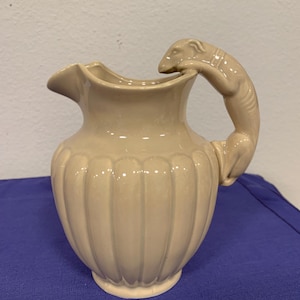 May include: Tan ceramic pitcher with a unique handle shaped like a squirrel. The pitcher has a ribbed design on the body and a spout for pouring. A decorative and functional piece for serving beverages.