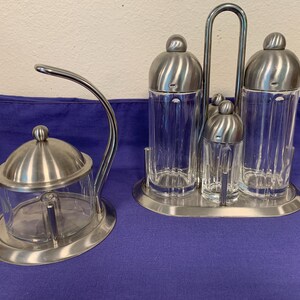 May include: A stainless steel and glass condiment set with a sugar bowl and a salt and pepper shaker set. The sugar bowl has a curved handle. The salt and pepper shakers are in a metal stand. The set is on a purple surface.