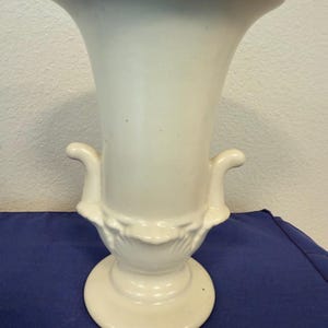 May include: Cream-colored ceramic vase with a flared top and a pedestal base. The vase features two curved handles and decorative floral detailing near the base. The vase is displayed on a blue surface.
