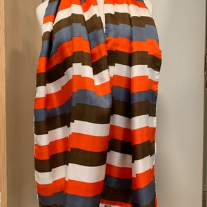 May include: A long, rectangular scarf with a geometric pattern of orange, white, brown, and blue stripes. The scarf is folded and draped over a mannequin.