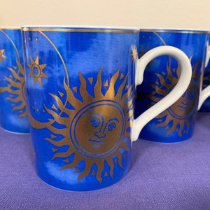 May include: Set of four blue ceramic mugs with gold sun and star designs. The mugs have white handles.