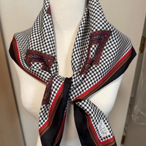 May include: A silk scarf with a black and white checkered pattern. The scarf has a red border with gold accents and a black border. The scarf is tied in a knot. The scarf has the text "SCOTTS & ANDREWS" on the corner.