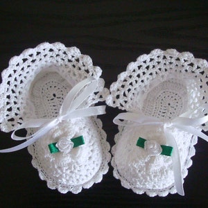 Puede incluir: Un par de botitas de bebé de ganchillo blanco con lazos de satén verde. Las botitas tienen un delicado borde de encaje y están atadas con cintas de satén blanco.