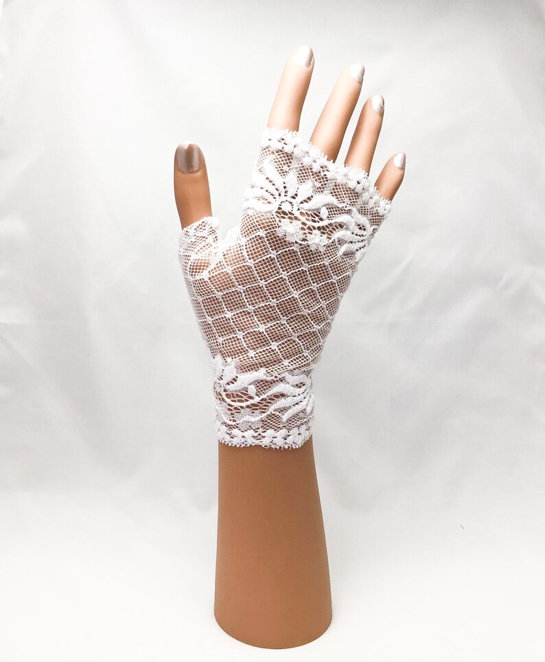 Puede incluir: Guantes sin dedos de encaje blanco con un delicado dise&ntilde;o floral y un dise&ntilde;o de red en forma de diamante. Los guantes se muestran en una mano de maniqu&iacute; sobre un fondo blanco. Un accesorio nupcial.