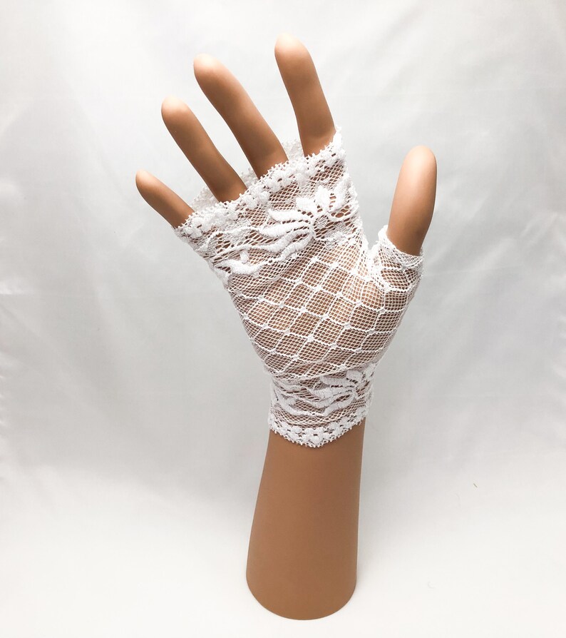 Puede incluir: Guantes sin dedos de encaje blanco con un delicado dise&ntilde;o floral en los bordes y un dise&ntilde;o de red en forma de diamante. Los guantes se muestran en una mano de maniqu&iacute; sobre un fondo blanco.