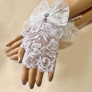 Puede incluir: Guantes sin dedos de encaje blanco con un estampado floral y un detalle de lazo. Los guantes están adornados con un pequeño adorno brillante en el centro del lazo. Los guantes están diseñados para ser usados en las manos.